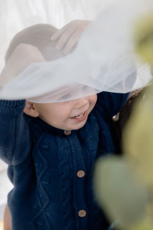 Smiling toddler in navy cable-knit sweater plays peek-a-boo under white tulle veil held by adult hands.
