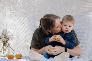 A loving parent kisses a toddler holding a lemon while sitting together against a white textured backdrop.