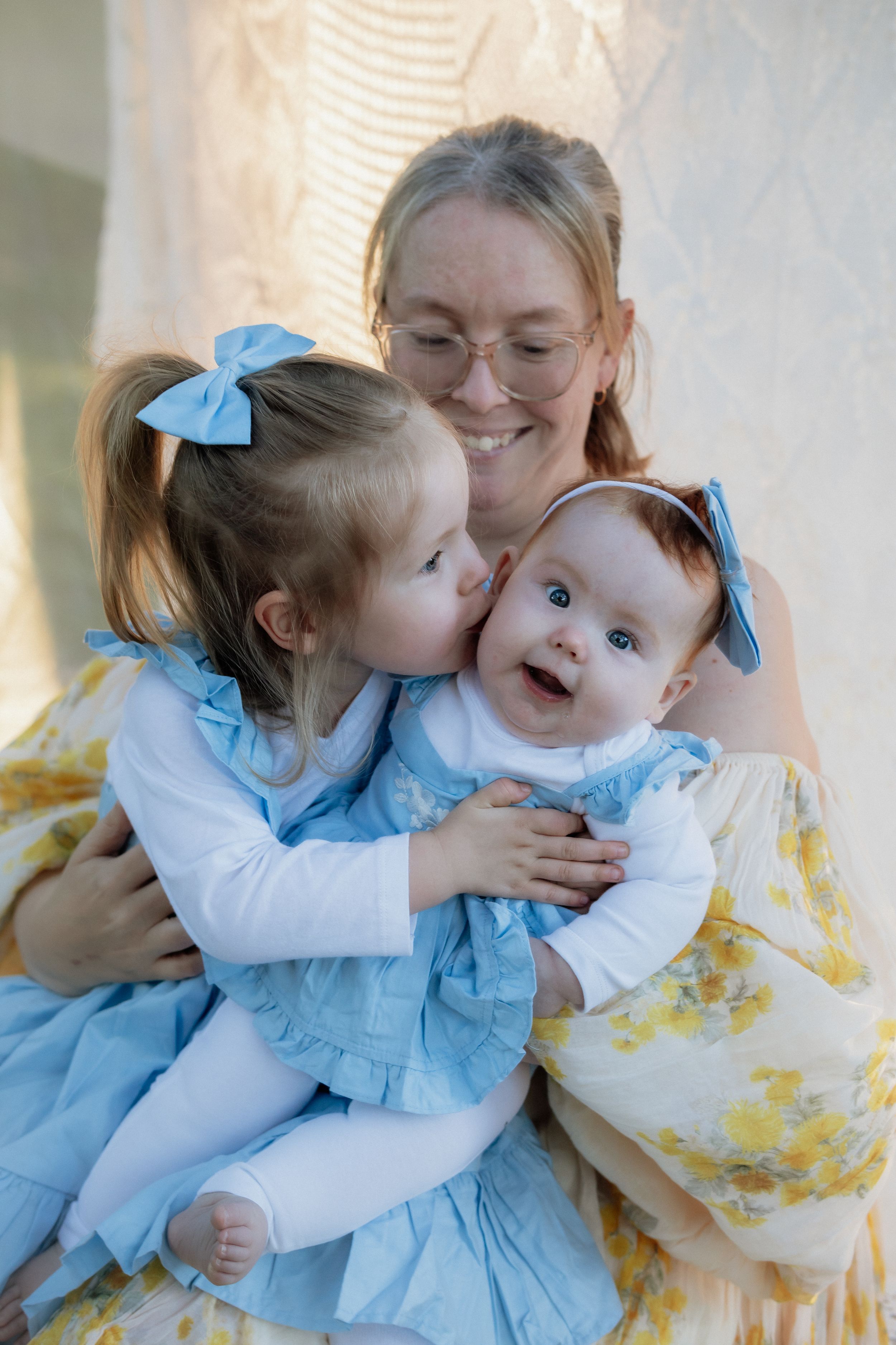 A smiling mother in a floral dress holds her baby and toddler daughter in blue dresses, sharing a motherhood moment.