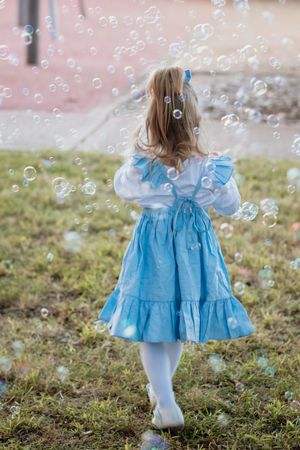 A young girl in a blue dress and white tights plays in a park in Kellyville surrounded by floating soap bubbles.