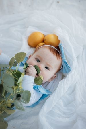 Baby in blue bow headband chewing green toy, lying on white fabric with yellow lemons and eucalyptus in a park in Kellyville.