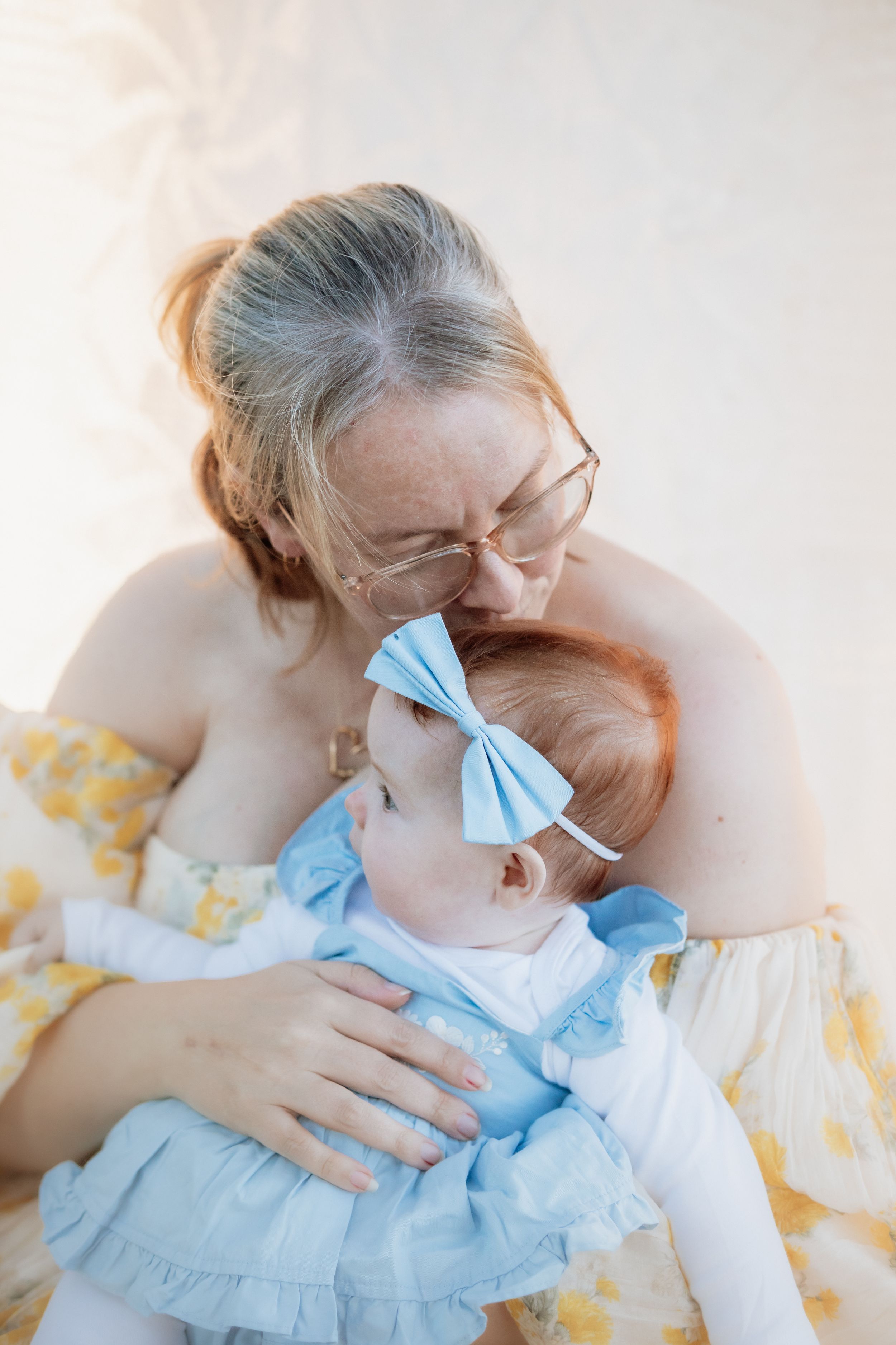 Mother with glasses kissing baby girl in blue dress and bow headband, sharing tender moment in a park in Kellyville.