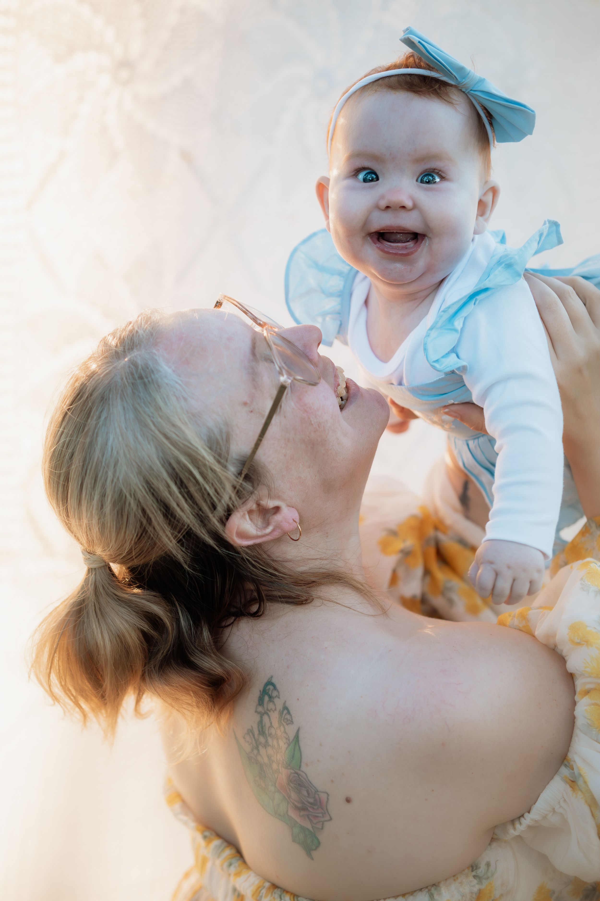 A joyful baby with blue eyes and bow headband being lifted by a tattooed woman in a bright, warm-toned setting in Beaumont Hills.