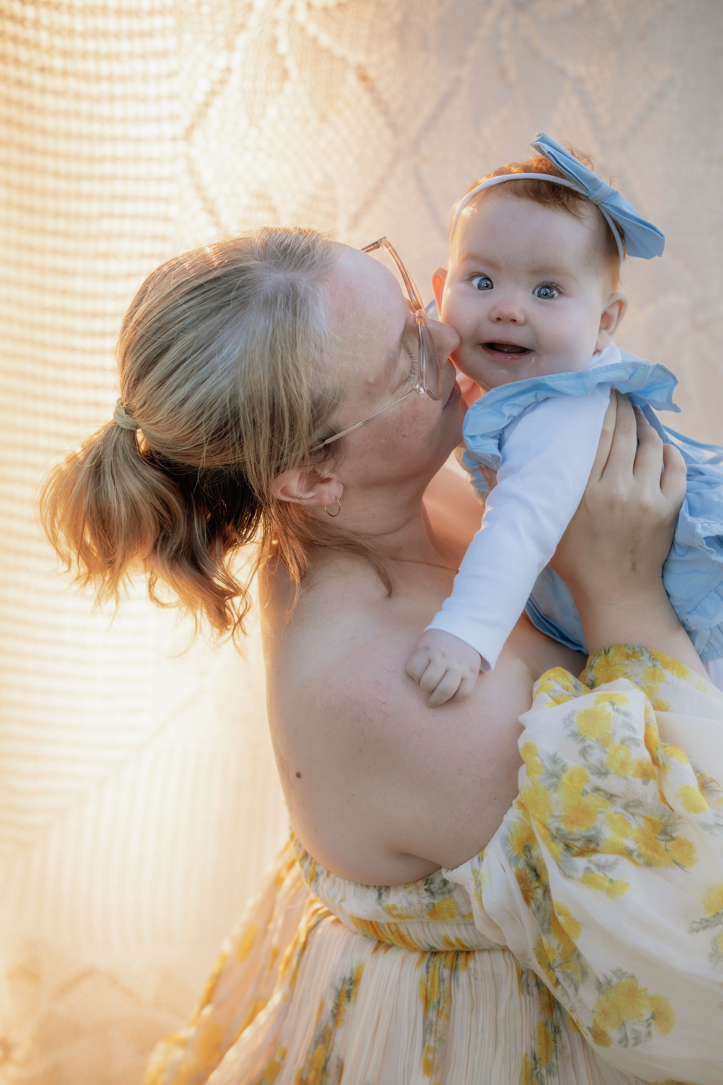 A Mother lovingly kisses her smiling baby in a blue bow and dress, bathed in warm golden light during mum and baby photos.