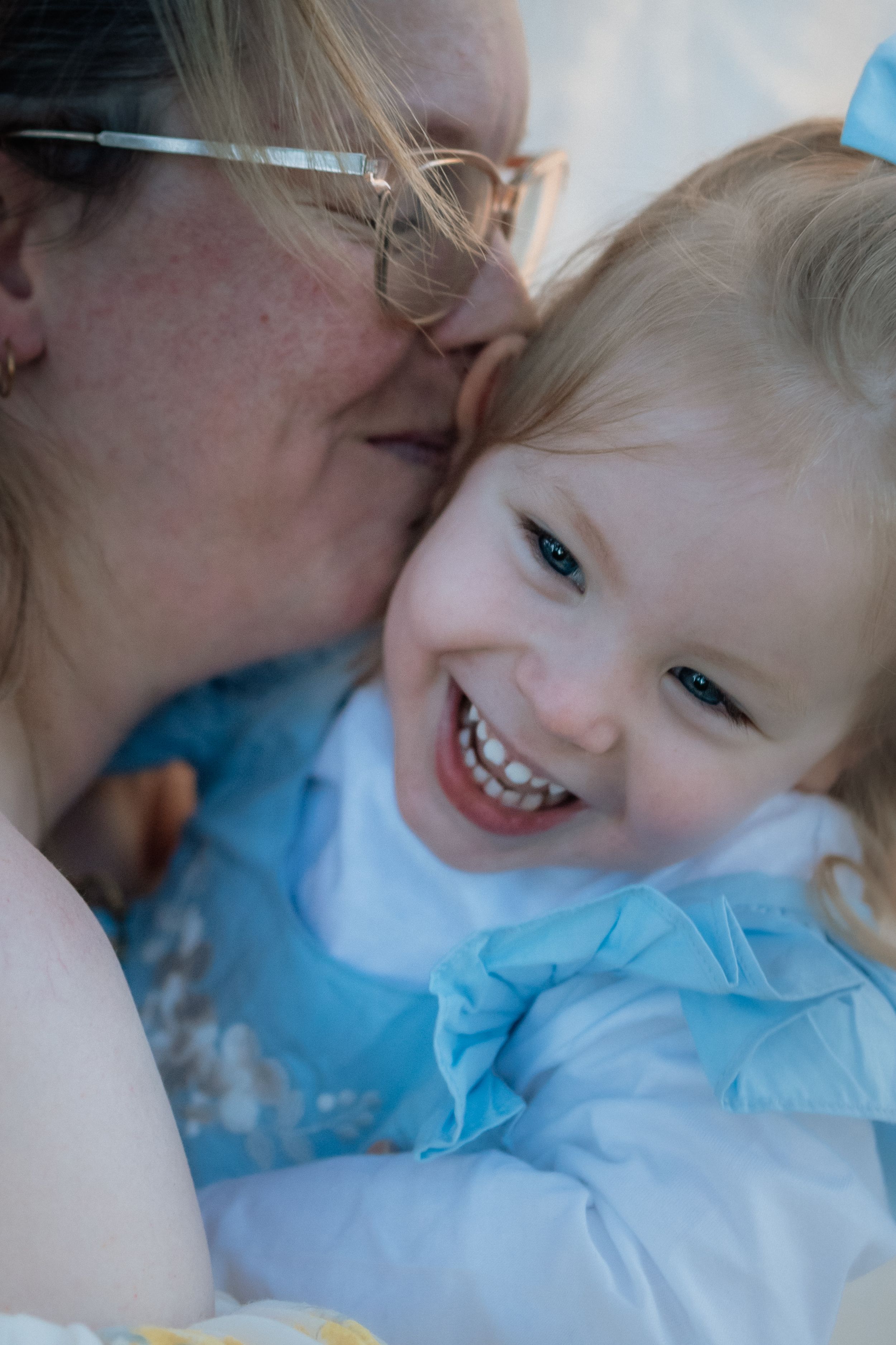 A woman with glasses kisses a laughing toddler girl in a blue dress, sharing a joyful tender motherhood moment.