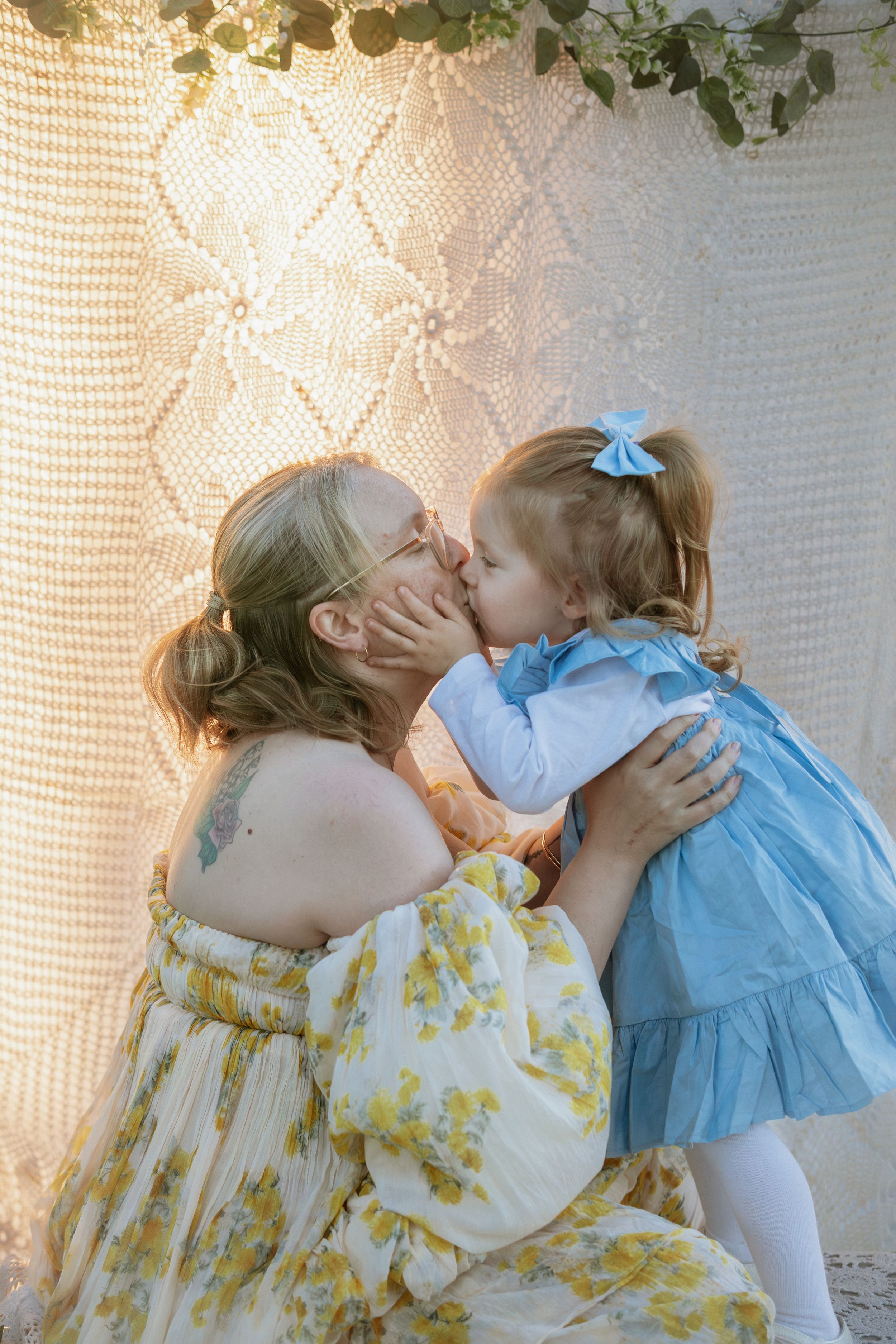 A mother in a floral dress receives a sweet kiss from her toddler daughter in a blue dress against a lace backdrop.