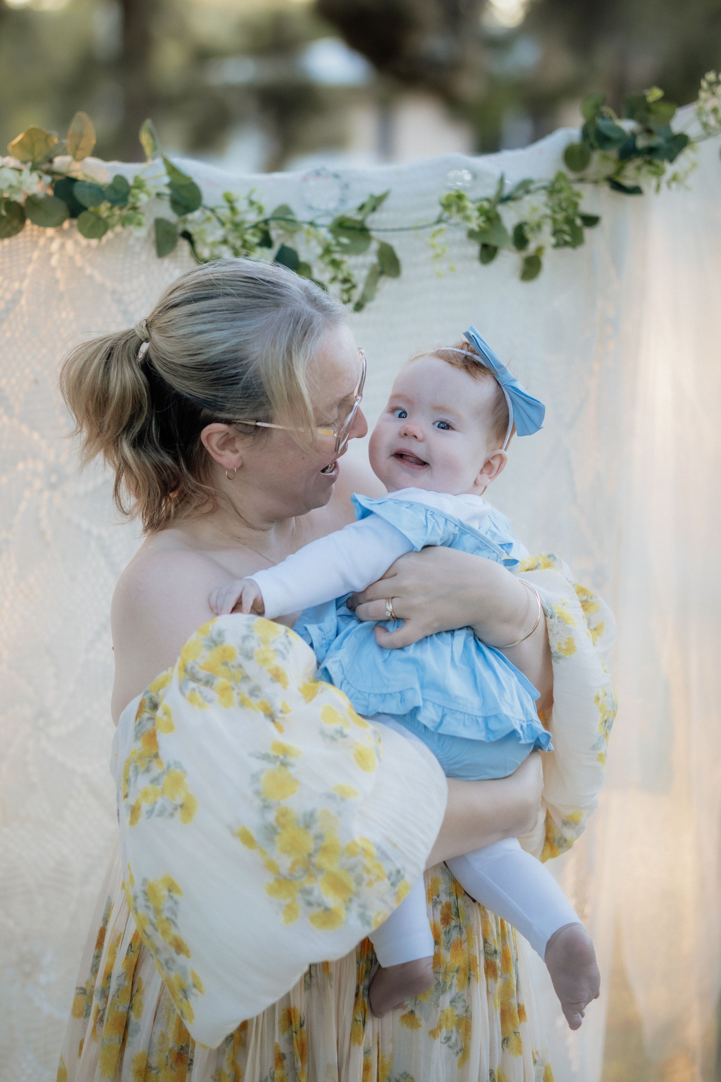 A smiling mother in a yellow floral dress lovingly holds her happy baby girl in a blue outfit outdoors.