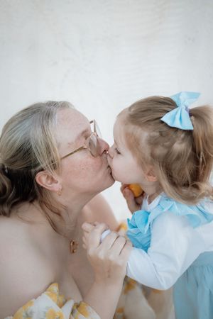 Mother with glasses sharing a kiss with a little girl wearing a blue bow, holding a fruit together during Mother and daughter photoshoot.