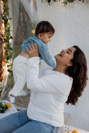 A joyful mum and baby photo in a white sweater lifts her smiling baby in a blue outfit outdoors near floral decor.