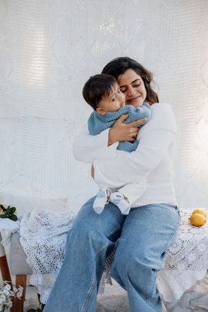 A smiling woman in white sweater and jeans lovingly embraces a toddler in blue knit sweater on a white lace bench in a park in Rouse Hill.