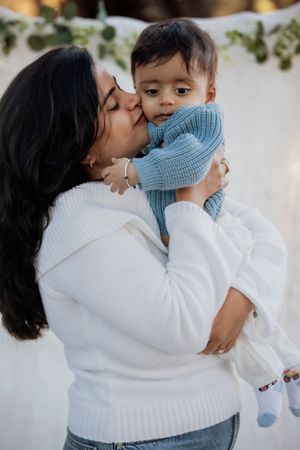 A mother in a white sweater kisses her baby dressed in a blue knit sweater, sharing a tender moment.