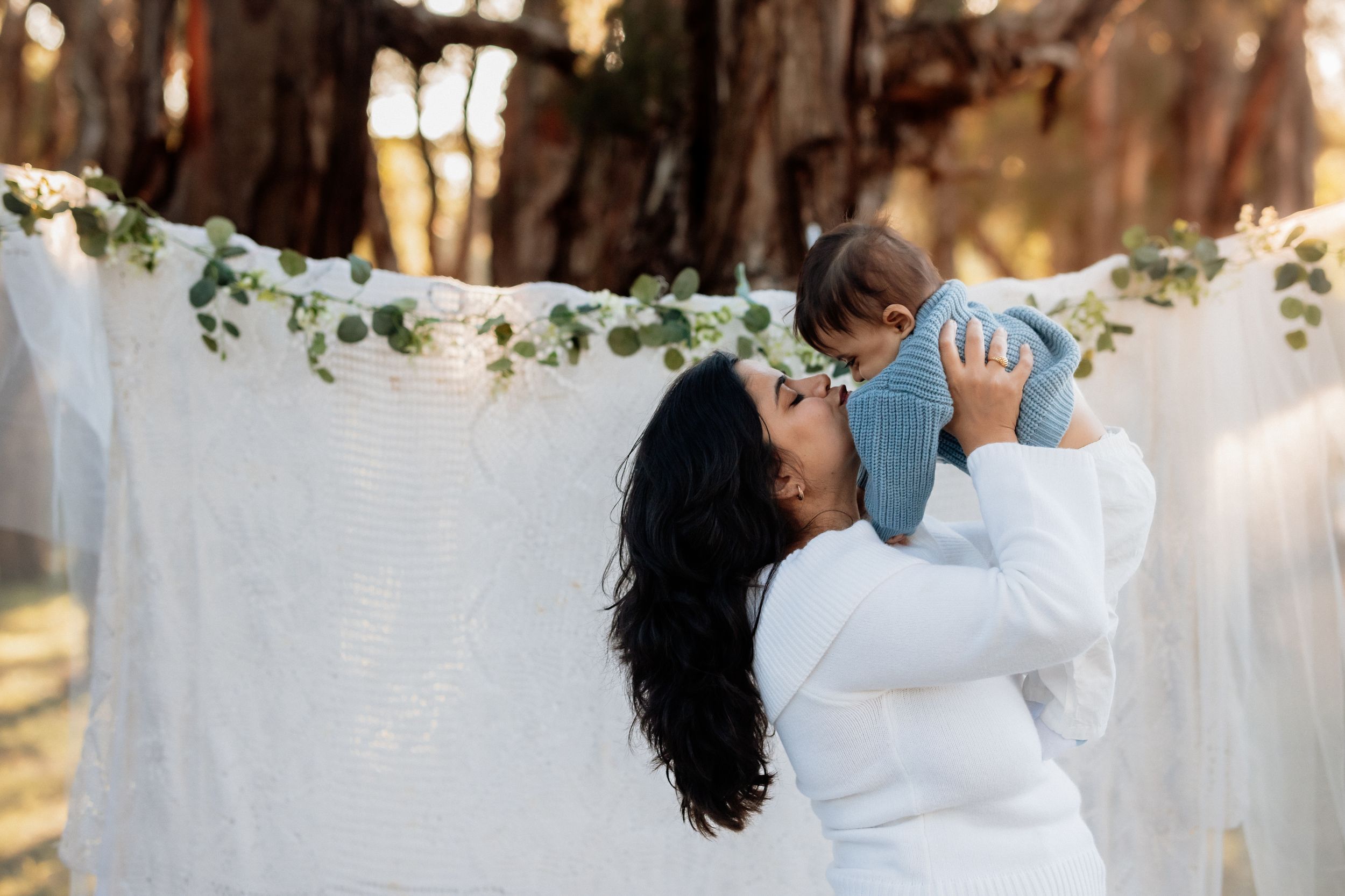 A mother in white lifts her baby outdoors against a flowing white backdrop with greenery garland in a park in Beaumont Hills.
