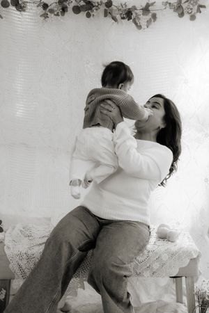 Black and white motherhood photo of a mother lifting and kissing her baby in a cozy decorated room.