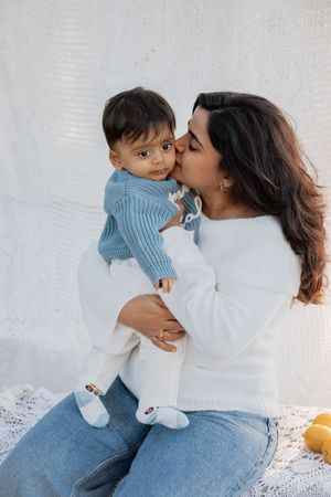 A woman in a white sweater kisses her baby dressed in a blue knit sweater and white pants in a park in Beaumont Hills.