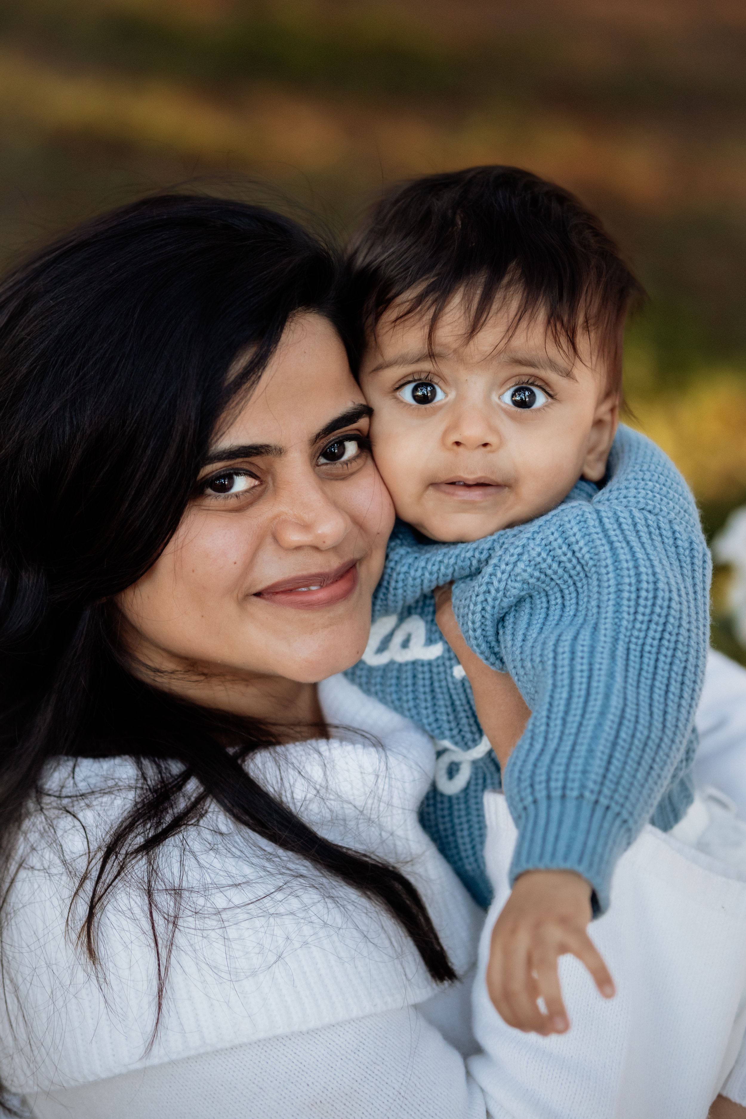 A smiling woman with dark hair holds a wide-eyed baby in a blue sweater outdoors in a park in Beaumont Hills.