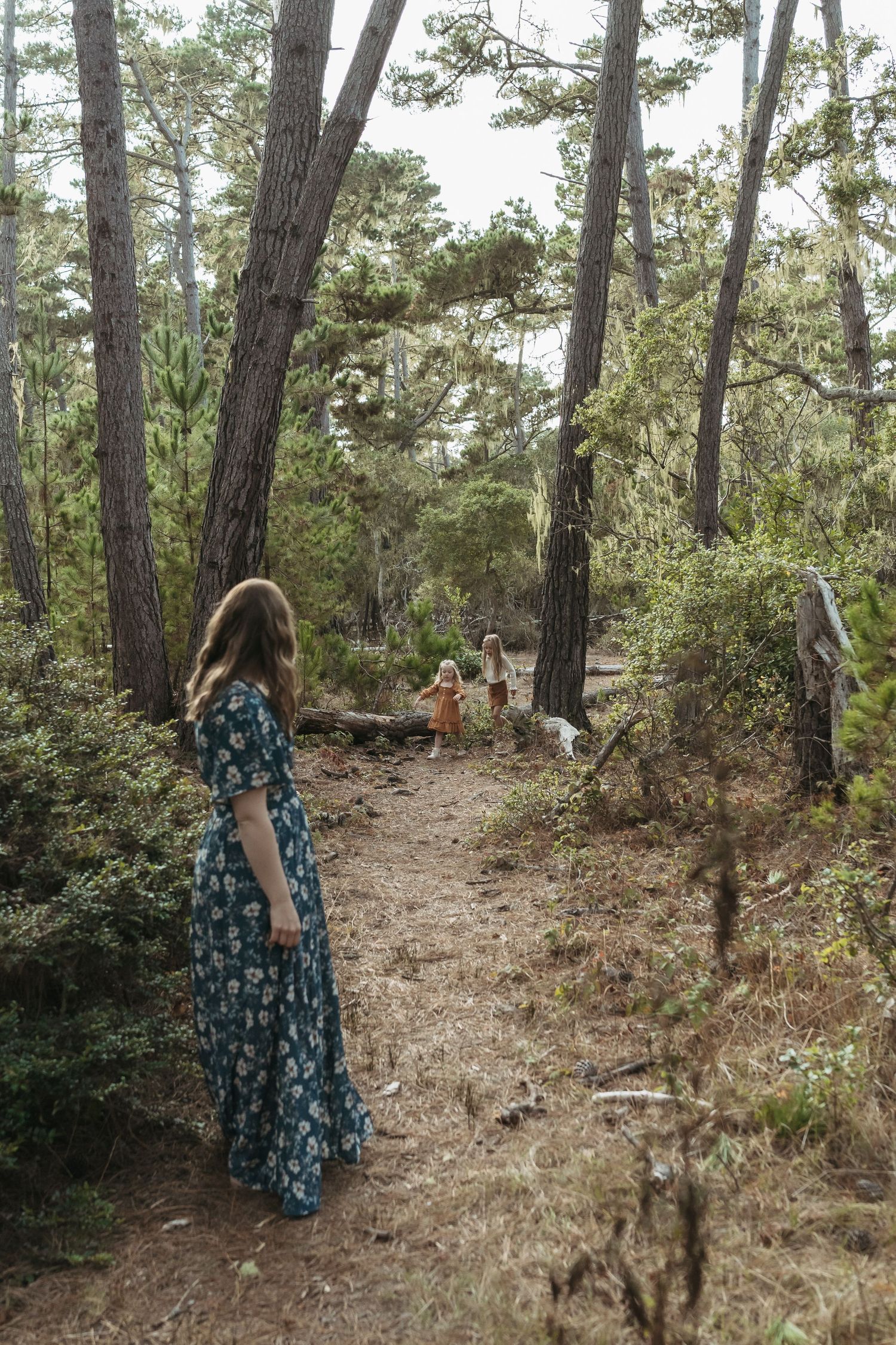 Woman in floral dress watches children walk along a forest path surrounded by tall pine trees.