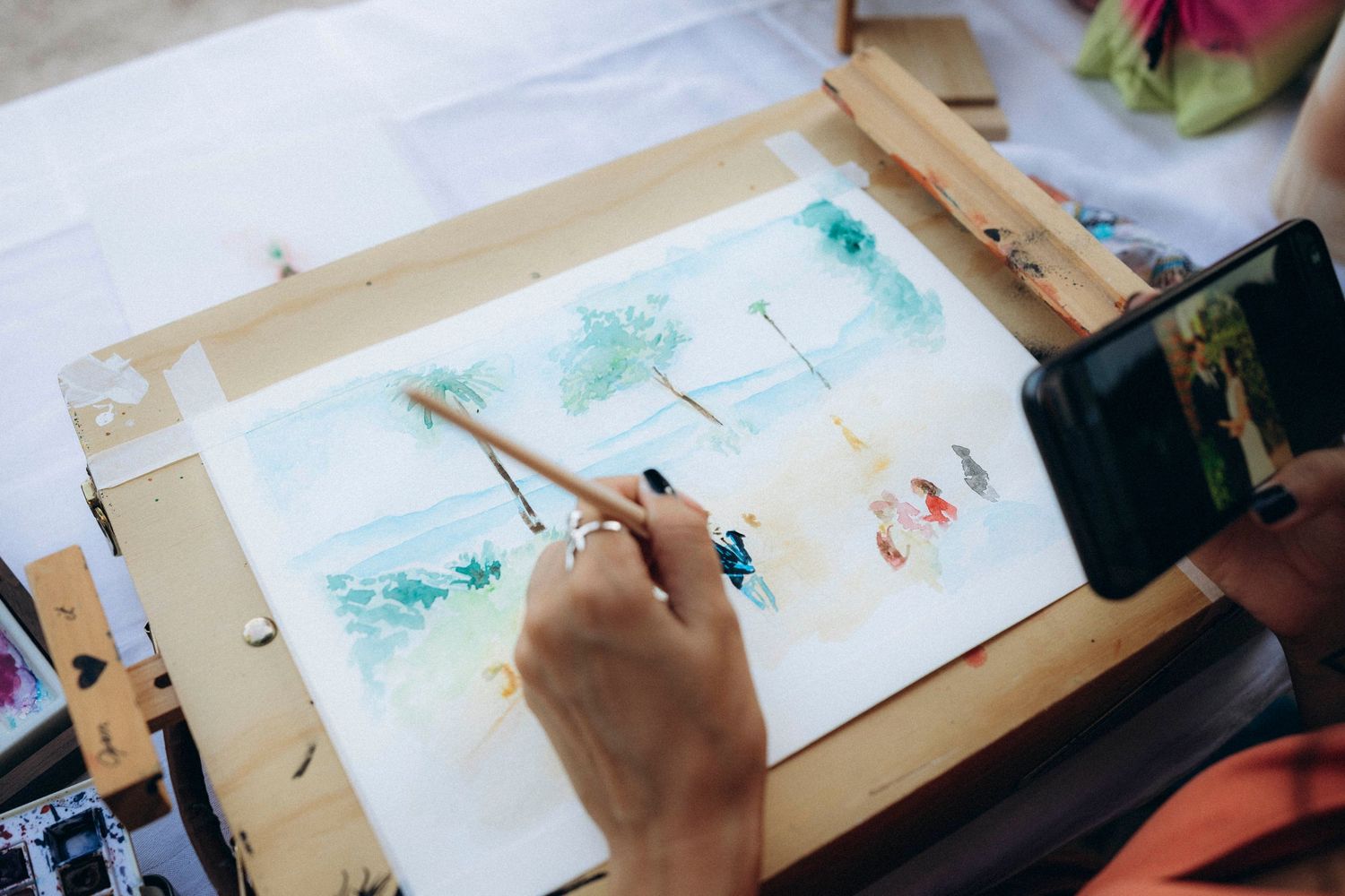 Artist painting a watercolor beach scene with palm trees on an easel while being filmed by a smartphone.