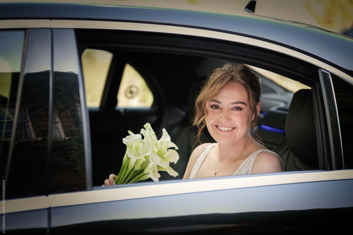 A smiling bride holds white calla lilies while sitting in a car on her wedding day.