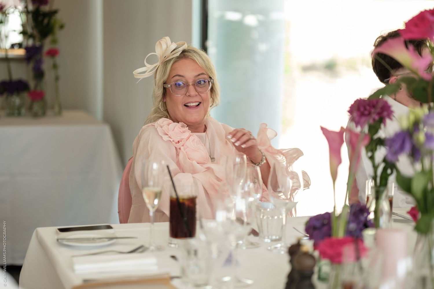 Elegant woman in pink outfit and fascinator hat smiling at a wedding reception table with floral arrangements.