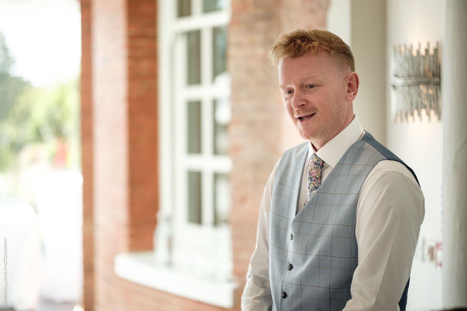 Smiling groom in light gray vest and floral tie at wedding venue.