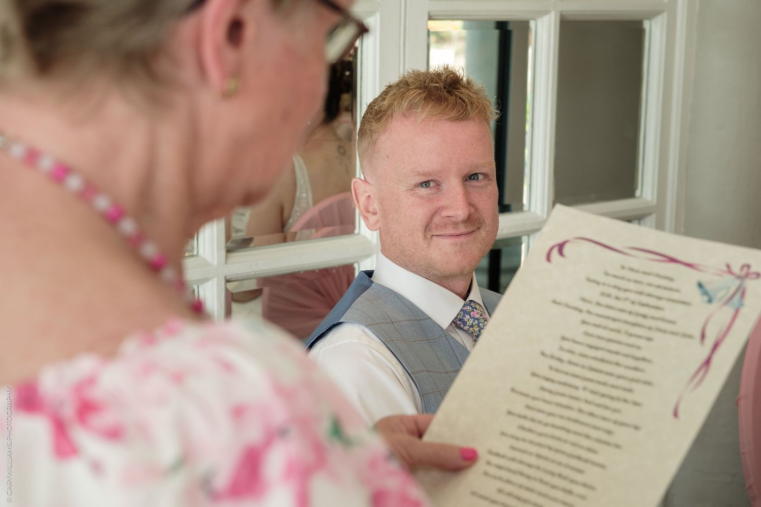 Groom smiling warmly at bride holding wedding ceremony script during indoor wedding celebration.