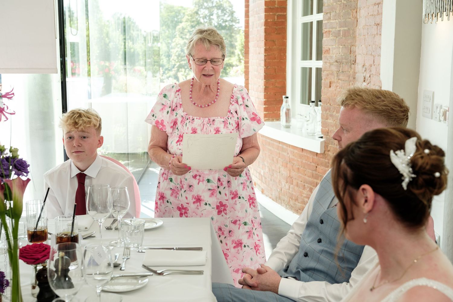 Elderly woman in floral dress giving a speech at a wedding reception dinner table with guests listening attentively.