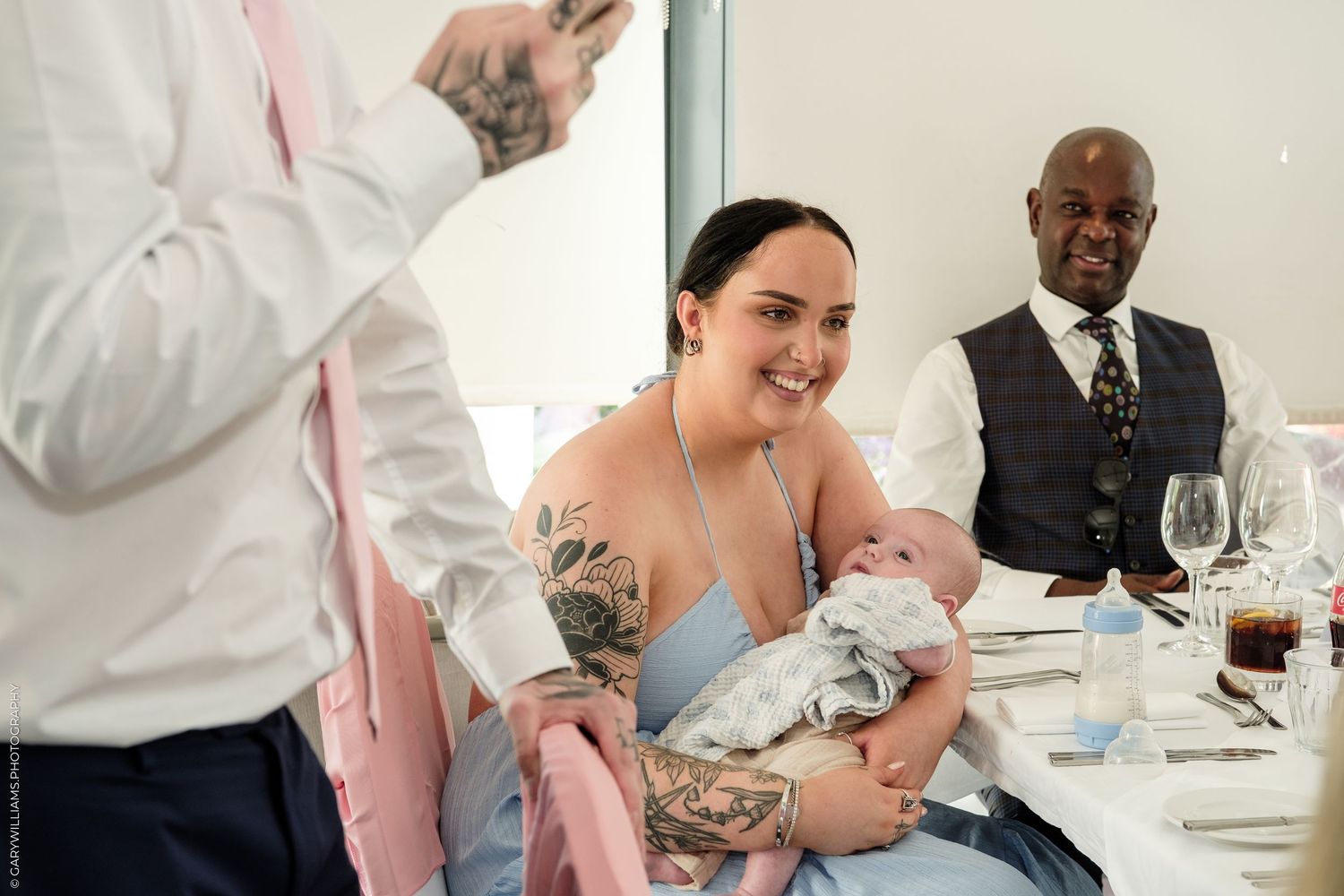Tattooed woman holding a baby at a wedding reception, smiling warmly while guests sit at an elegant table.