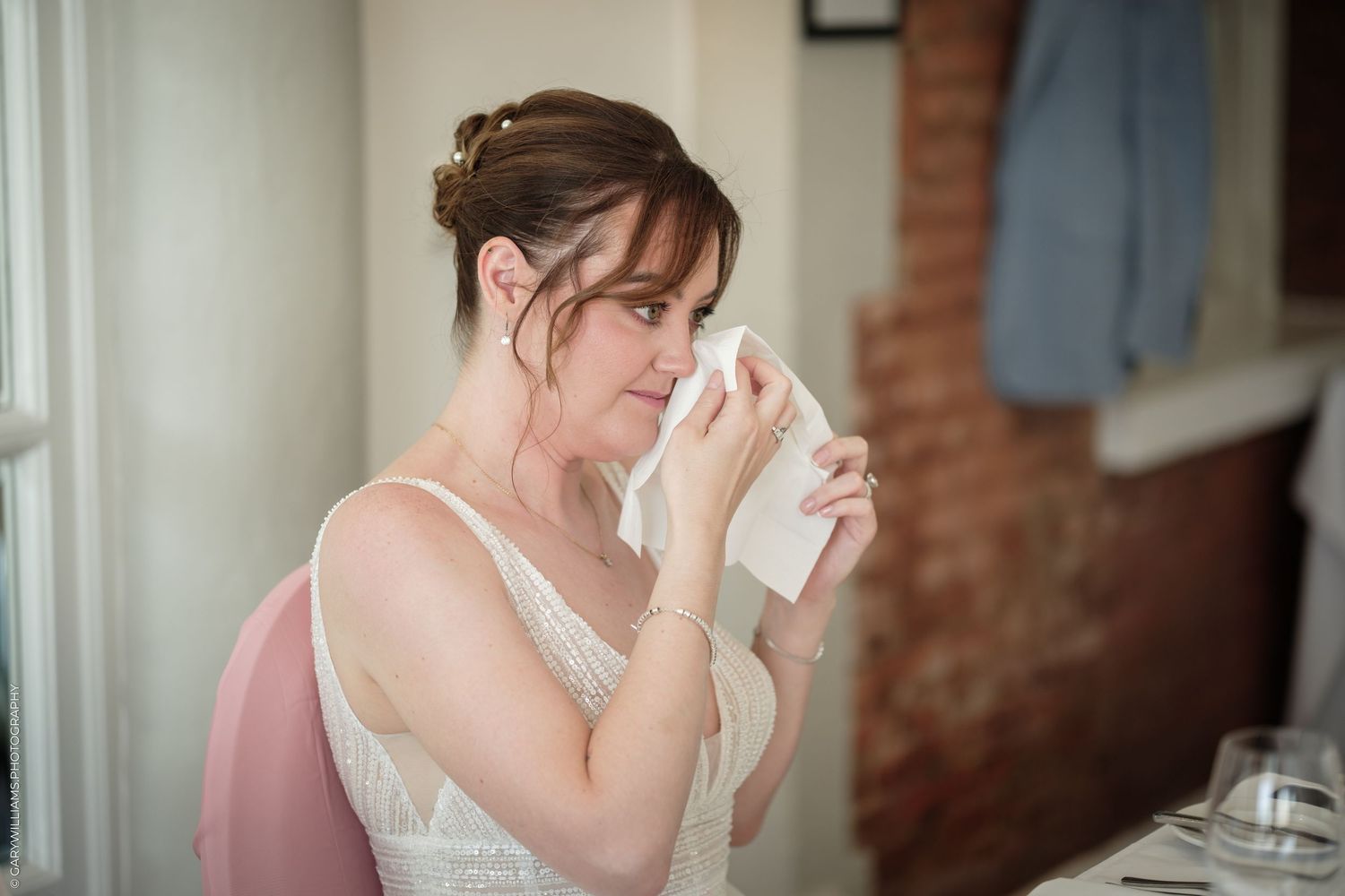 Emotional bride in white lace dress dabbing tears with tissue during wedding preparations in vintage venue.