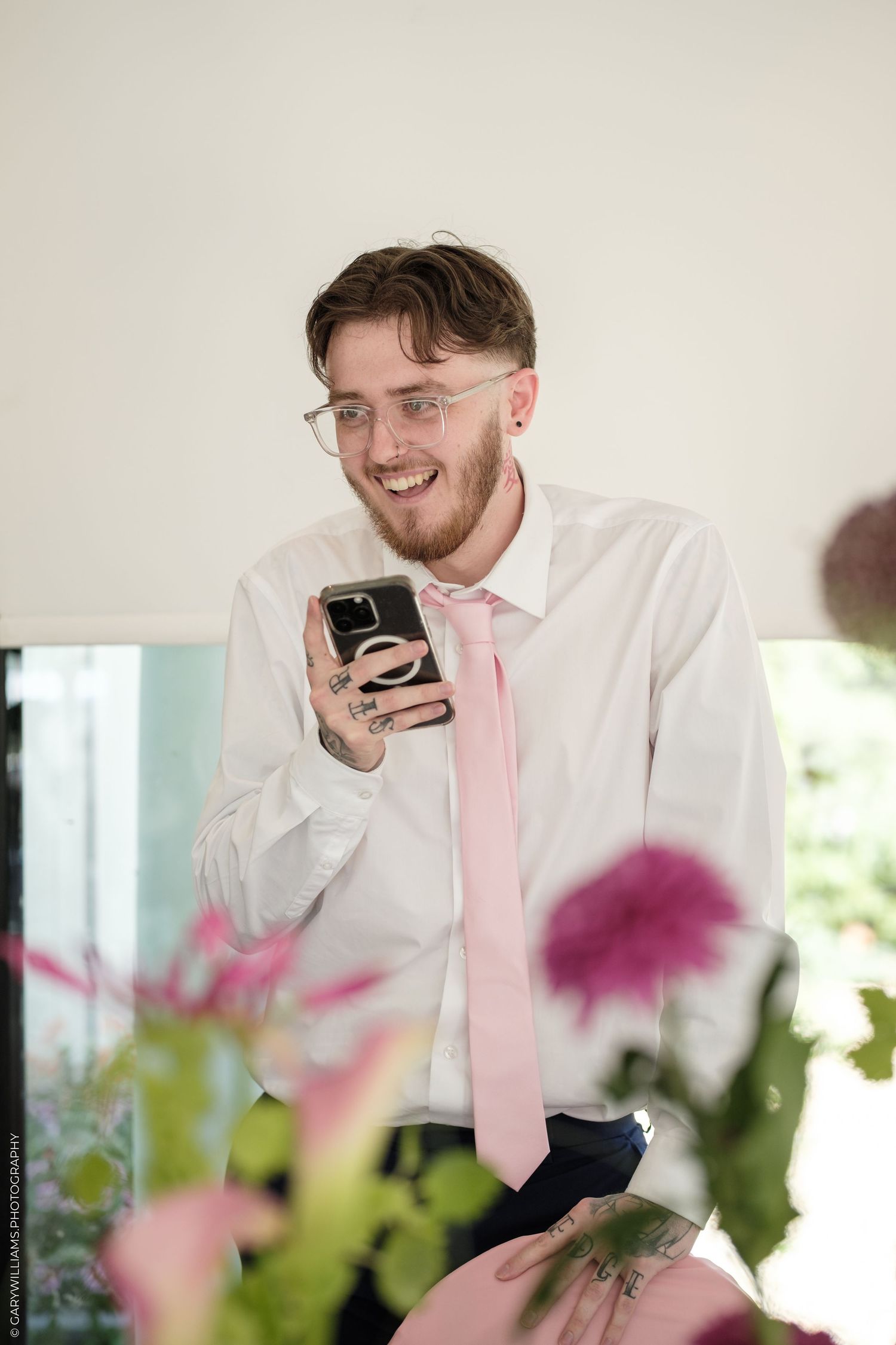 Smiling man with glasses and pink tie holding smartphone, surrounded by pink flowers in bright setting.