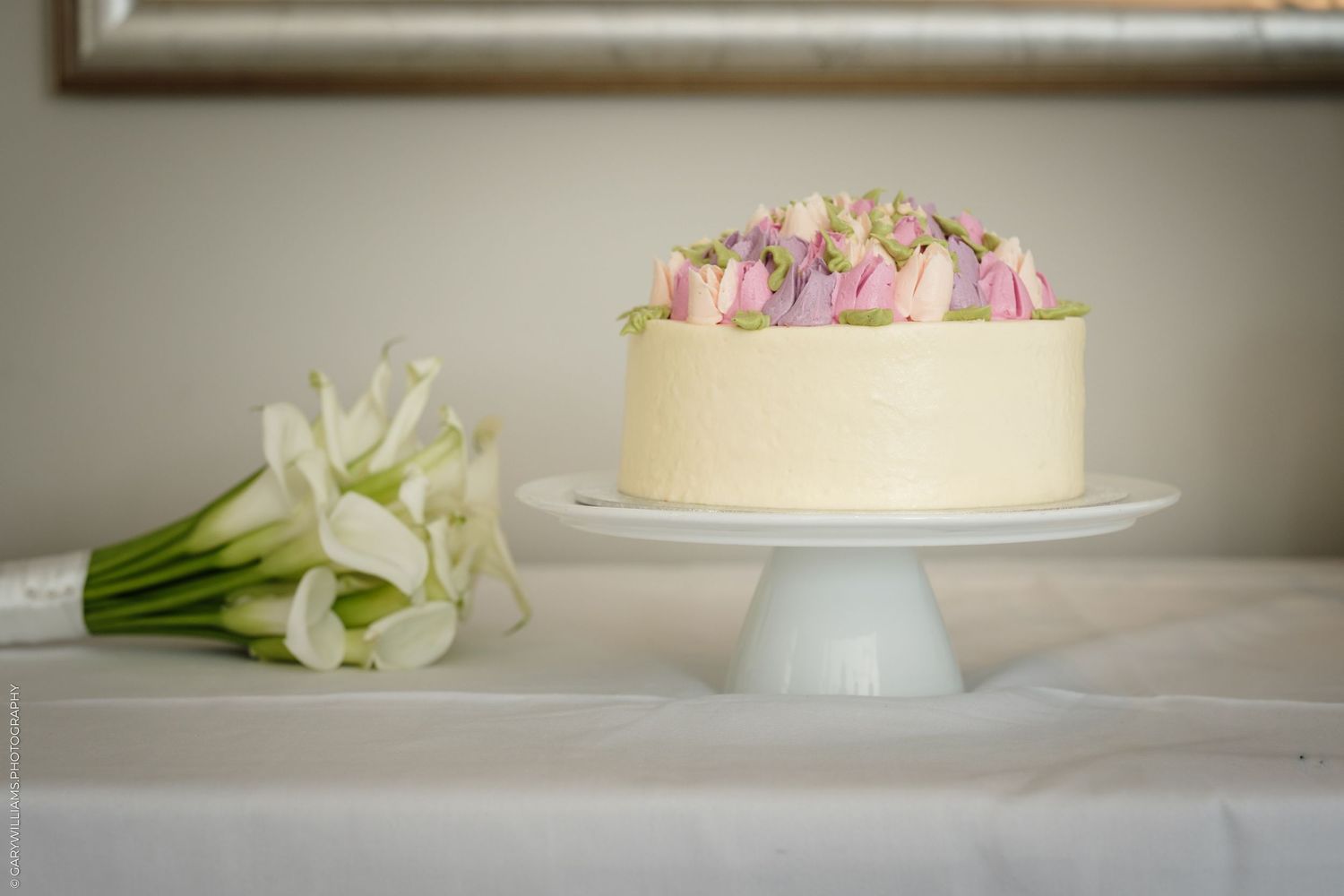 Elegant white cake with pink roses on a cake stand beside white calla lilies bouquet on a draped table.