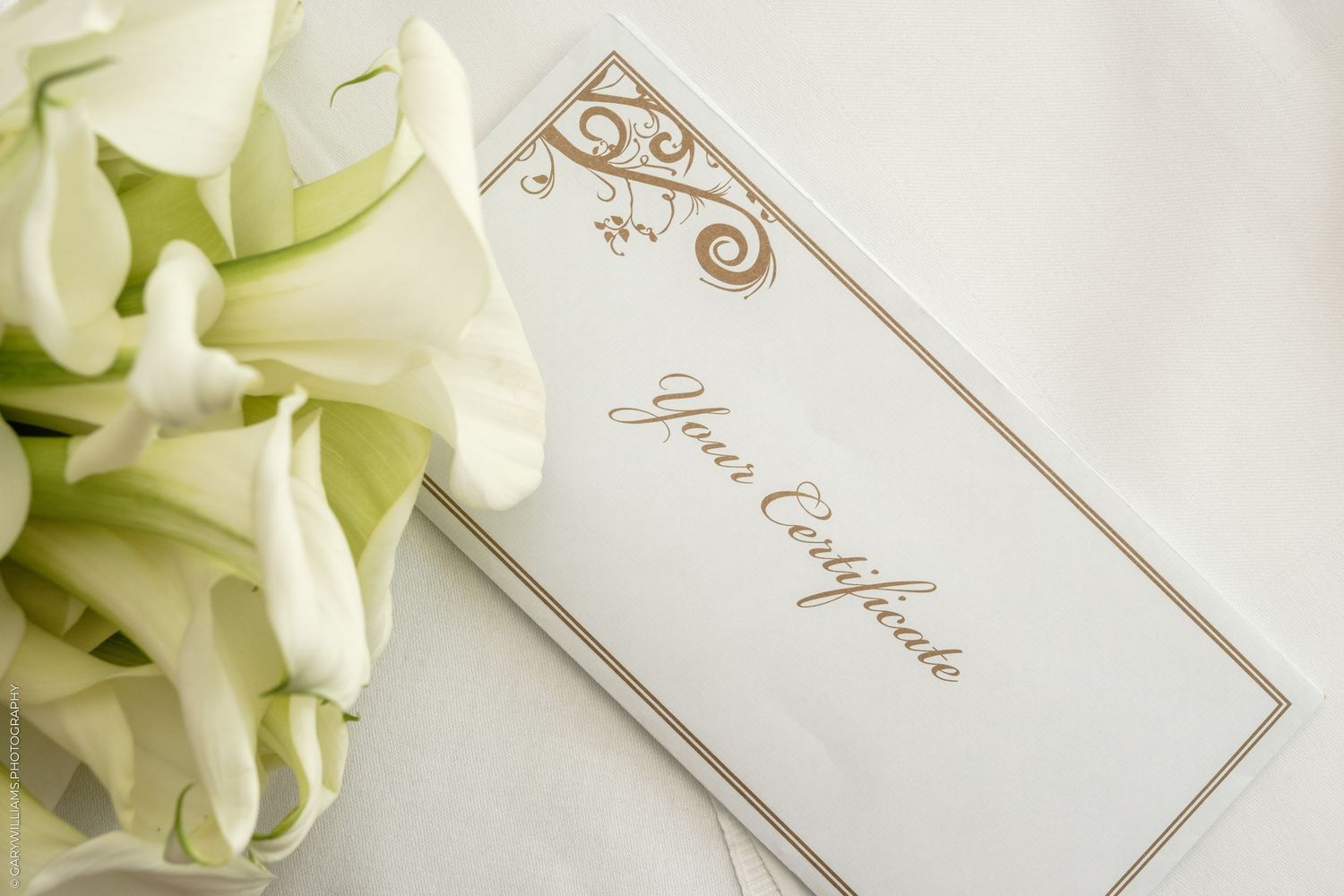 Elegant gift certificate with gold script beside white calla lilies on soft white fabric background.