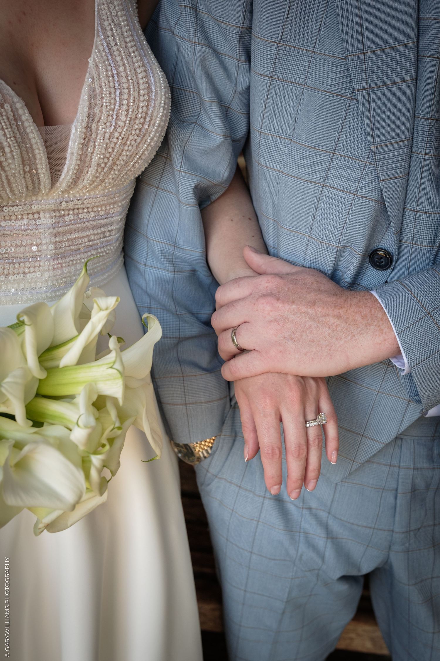 Newlyweds display wedding rings, bride holding white calla lily bouquet, groom in light blue plaid suit.