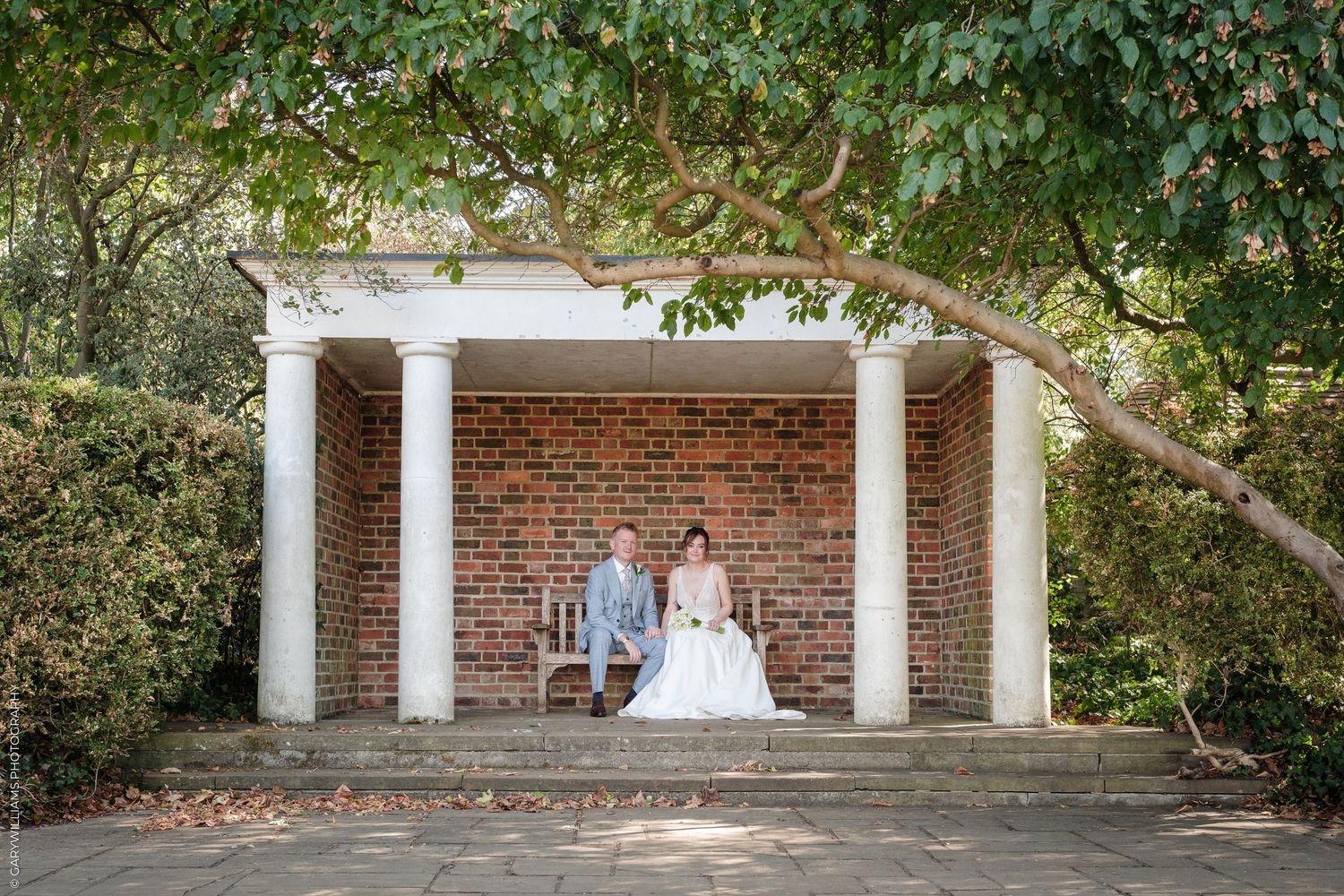 Bride and groom sitting under a brick columned gazebo surrounded by lush greenery on their wedding day.
