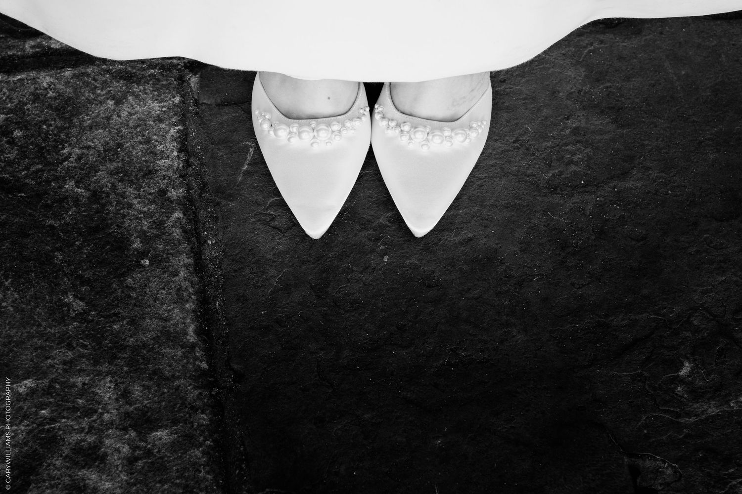 Black and white photo of white bridal heels with pearl details peeking beneath a wedding dress on stone floor.