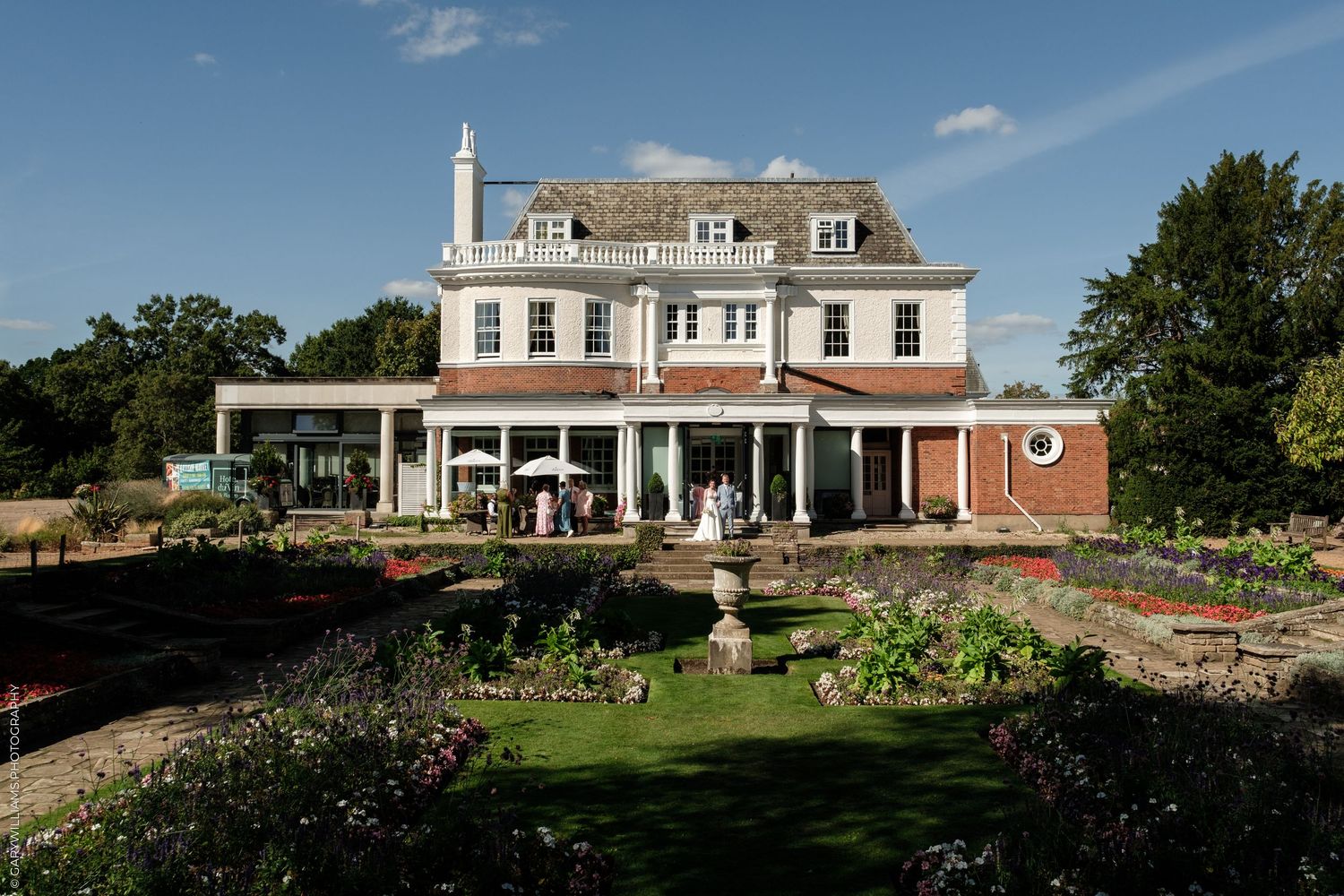 Elegant Georgian country house wedding venue with manicured gardens, red brick accents, and guests gathered on the lawn.