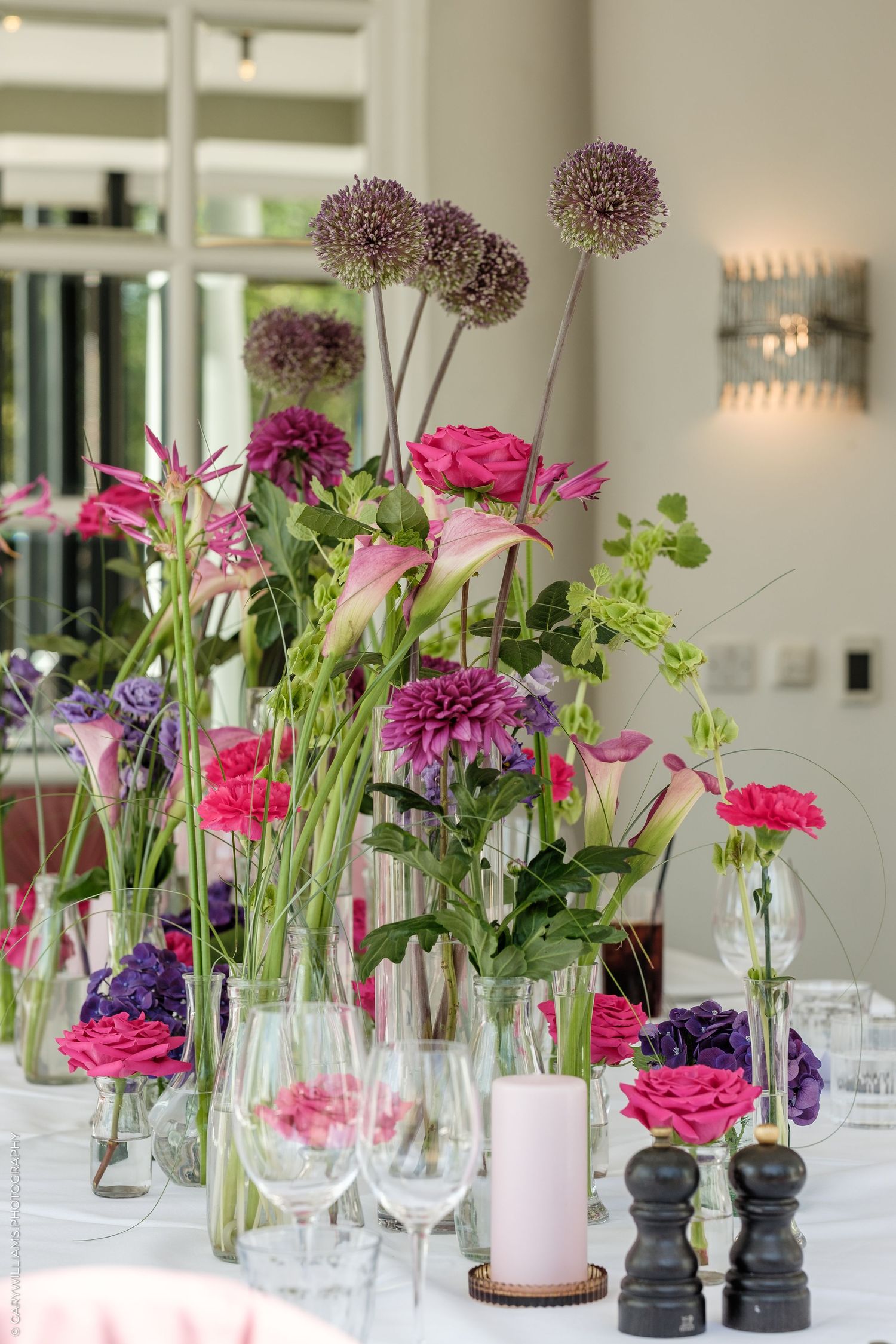 Elegant table centerpiece with pink roses, purple alliums, and dahlias in glass vases beside wine glasses and candles.