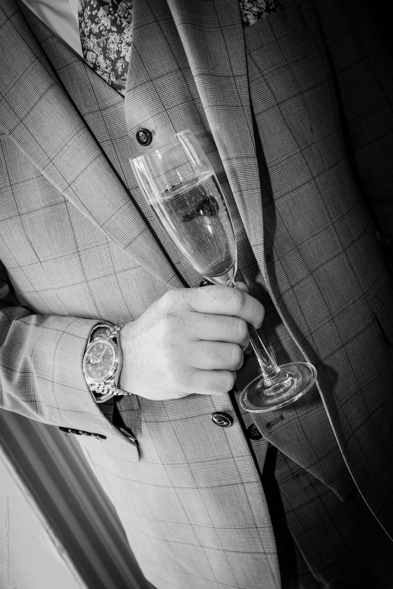 Black and white photo of a man in a plaid suit holding a champagne flute, wearing a luxury wristwatch.