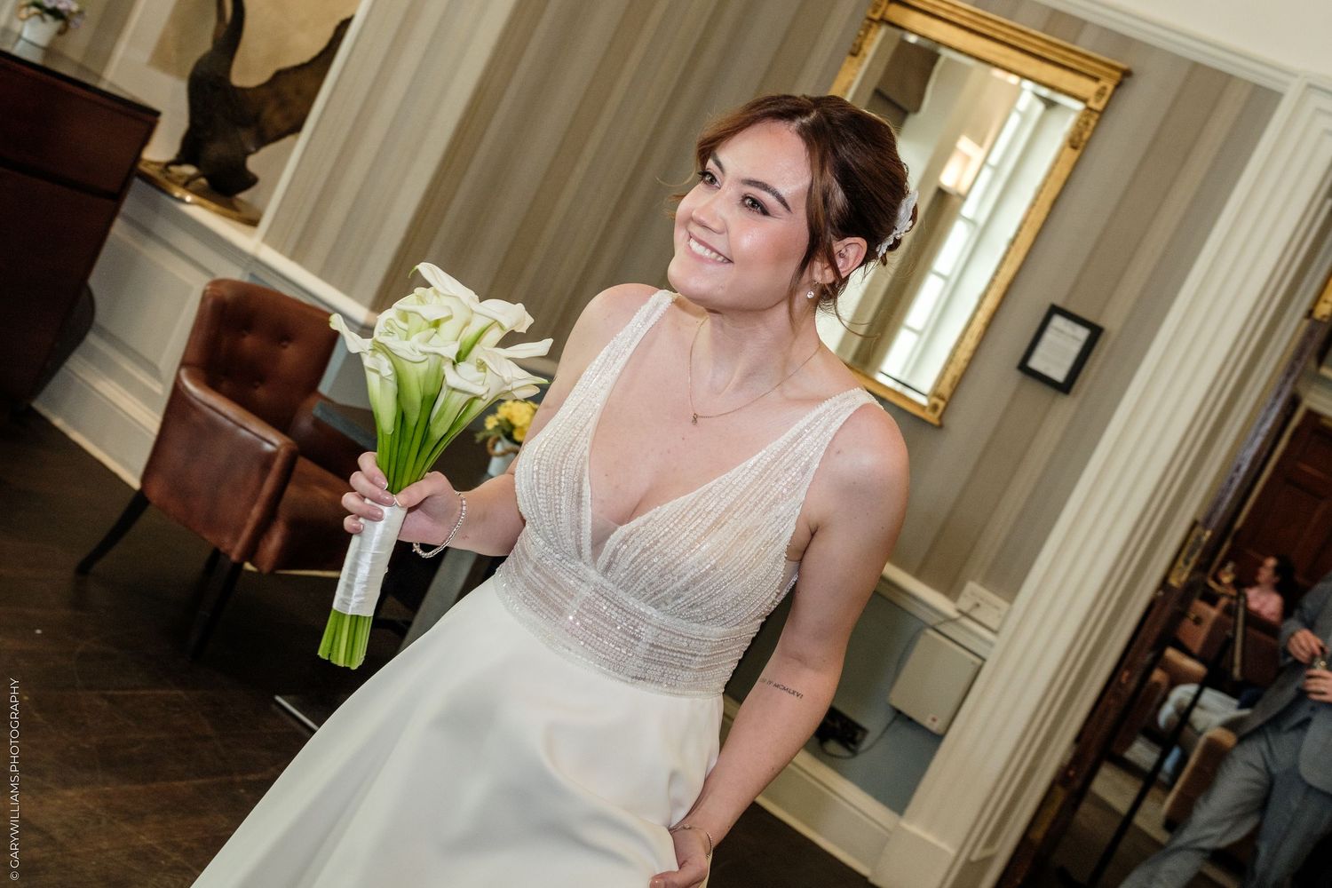 Smiling bride in white V-neck gown holding white calla lily bouquet in elegant room with mirror.