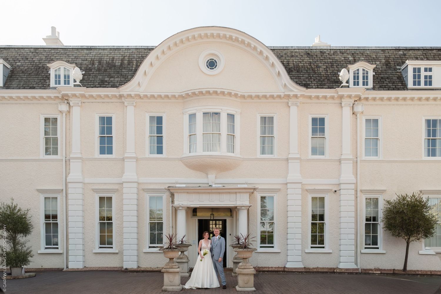 Bride and groom pose before elegant white Georgian manor house wedding venue with grand entrance and manicured grounds.
