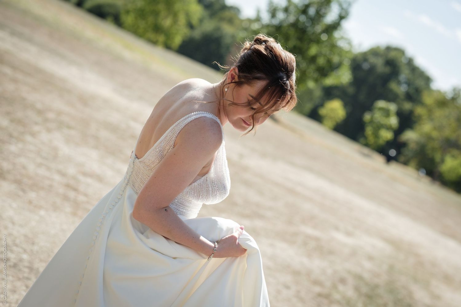 Bride in white gown smiling outdoors in sunlit field with green trees in background.