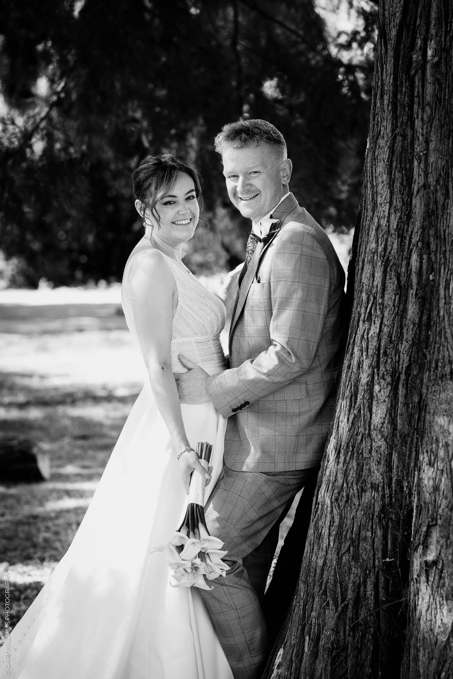 Black and white wedding photo of smiling bride and groom leaning against a tree outdoors.