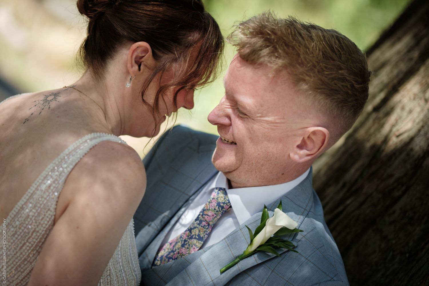 Bride and groom share an intimate moment, smiling at each other, groom wearing a blue suit with floral tie and boutonniere.