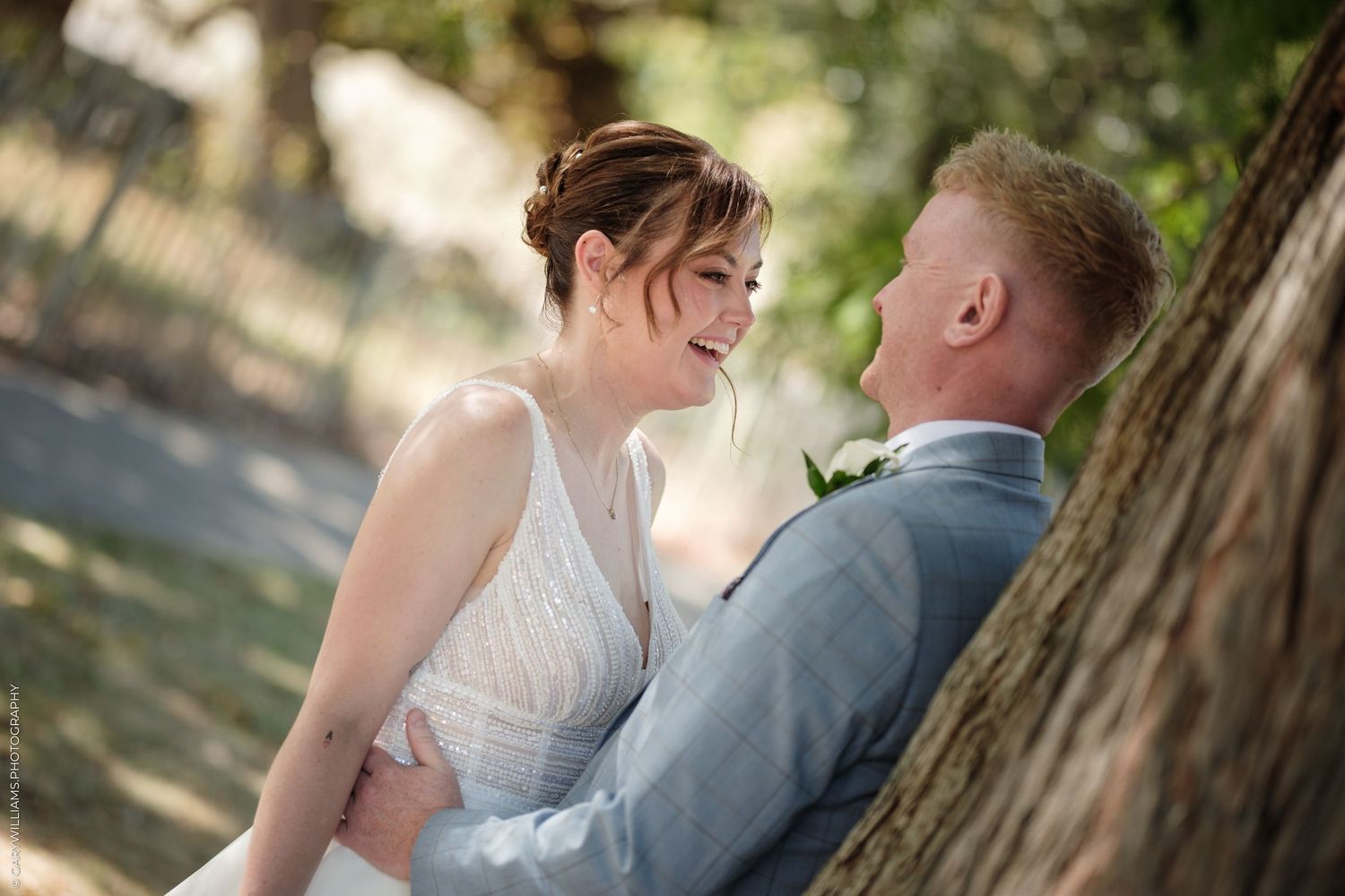 A smiling bride in a white dress gazes at her groom in a gray suit, leaning against a tree outdoors.