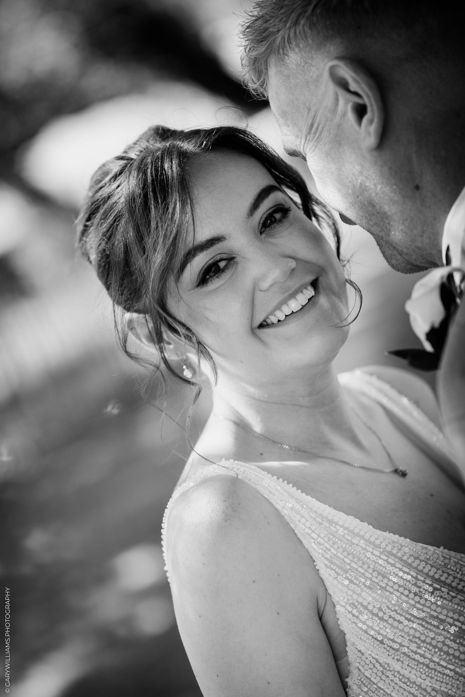 Black and white wedding photo of a smiling bride looking at camera while groom leans in close.