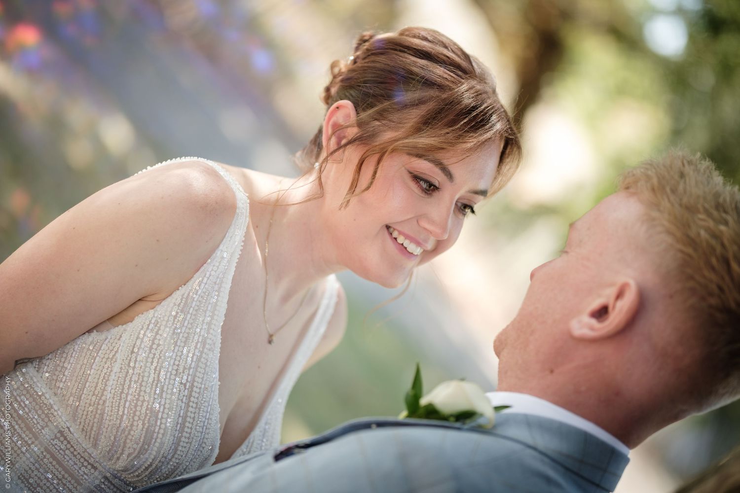 A bride in a silver gown smiles lovingly at a man in a suit during an outdoor wedding ceremony.