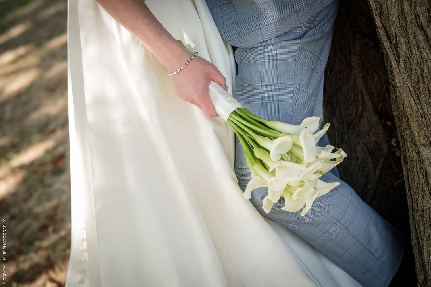 Bride holding white calla lily bouquet in a wedding gown beside groom in light blue suit near a tree.