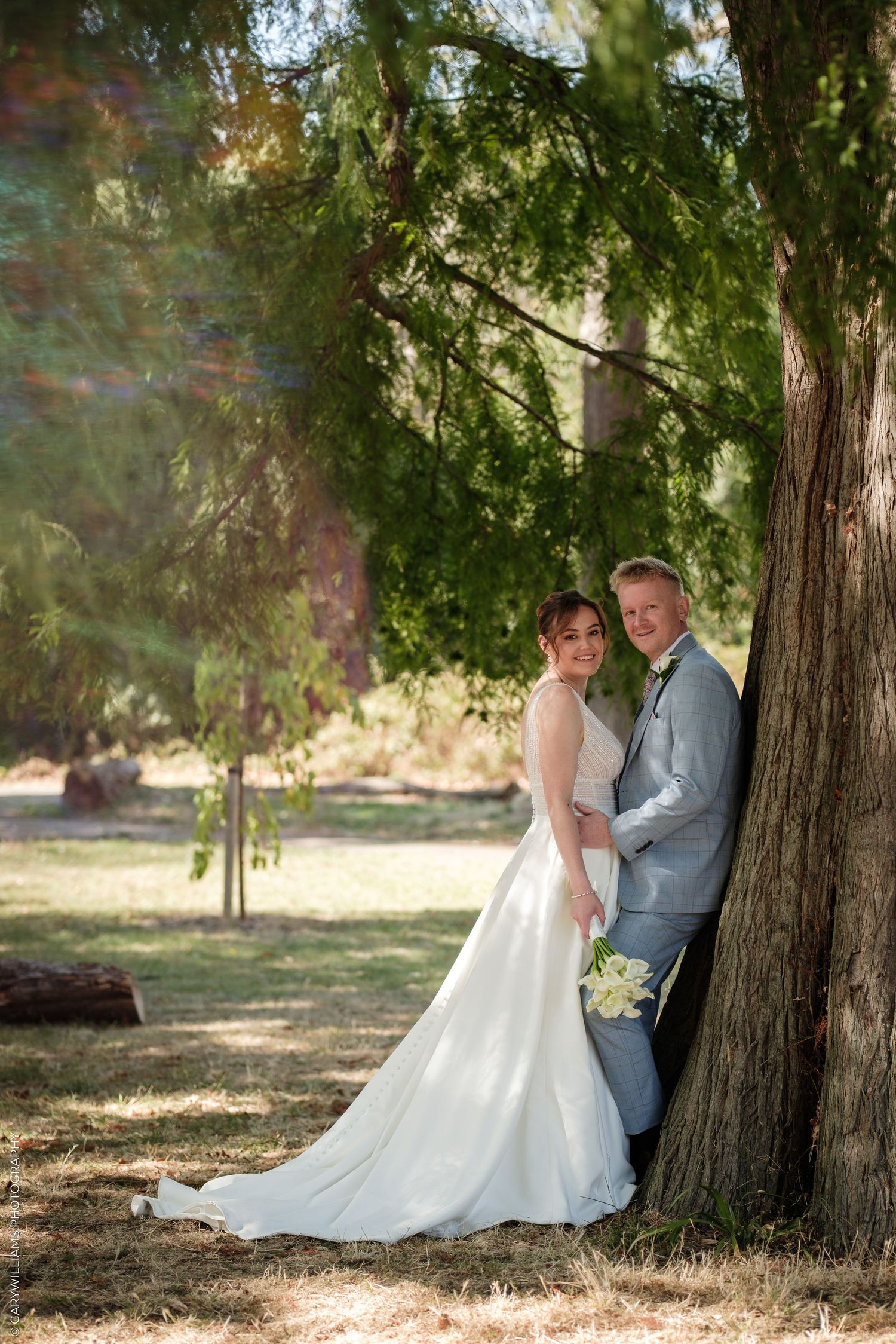 Bride and groom leaning against a large tree in a sunlit park, bride holding white flowers in a elegant white gown.