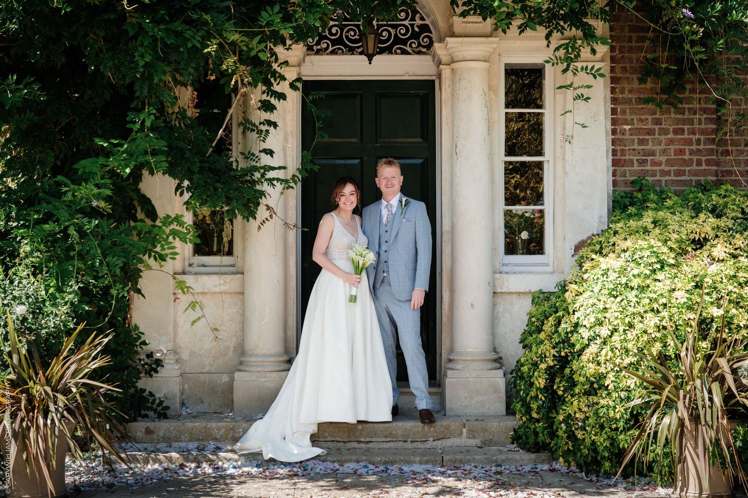 Bride and groom posing together at elegant manor house entrance, surrounded by lush greenery on their wedding day.
