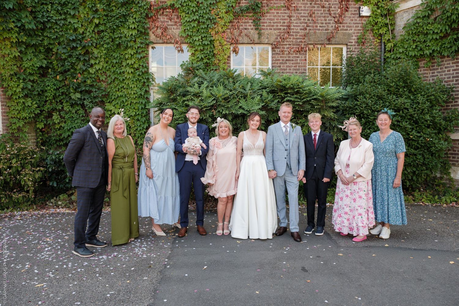 Wedding party posing together outdoors in front of ivy-covered brick wall, bride in white dress with bouquet.