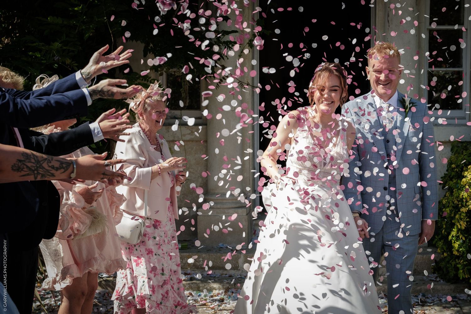 Bride and groom celebrate with confetti shower as guests toss pink flower petals outside wedding venue.